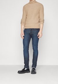 Beige ribbad tröja med hög hals, mörkblå slim-fit jeans, och svarta slip-on skor med kraftiga sulor. Neutral bakgrund.