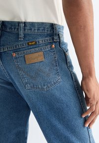 Nærbilde av en person som har på seg blå Wrangler-jeans med synlig baklomme, lærlogo-merking, og hånd hvilende ved siden av.