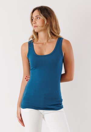 Top aderente blu con scollatura a barca e spalline larghe, realizzato in morbido tessuto elasticizzato. Indossato con pantaloni bianchi attillati.
