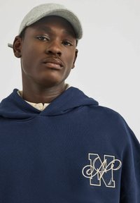 Felpa con cappuccio navy dal texture morbida e logo ricamato color crema sul petto. Completata da un cappellino grigio chiaro.