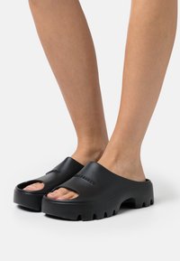 Musta kumiset liukuhäntäklotsit avoimella varpaalla ja paksulla pohjalla, joissa on sileä pinta ja kohokuvioitu logo hihnassa.