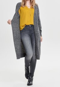 Vrouw die een mosterdgele top draagt, een lange grijze cardigan, verweerde zwarte skinny jeans en zwarte enkellaarsjes, lopend naar voren op een witte achtergrond.