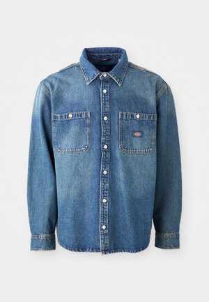 Denim shirt in medium blue, featuring a pointed collar, two chest pockets, and silver buttons. Long sleeves with buttoned cuffs.