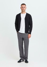 Young man standing with hands in pockets wearing black cardigan, white t-shirt, gray pants, and black sneakers with white stripes.