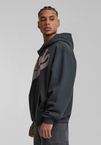 Mørkegrå hoodie med lynlås foran, prydet med et tekstureret grafisk design i sølv og rød på brystet. Inkluderer kengurulommer.