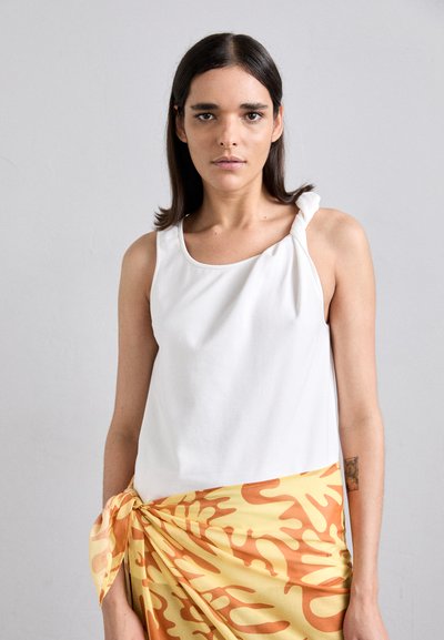 White sleeveless top with a knotted shoulder detail, paired with a yellow skirt featuring an abstract orange pattern. Smooth texture.