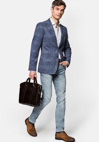 Dopasowana niebieska marynarka w kratę, biała koszula, jasne dżinsy, brązowe buty. Trzyma ciemno-brązową skórzaną teczkę z uchwytami na górze.