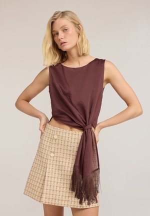 Femme blonde portant un haut marron sans manches noué à la taille avec des franges et une jupe beige à carreaux boutonnée, posant avec les mains sur les hanches.