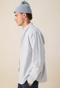 Jonge man in een lichtblauwe gebreide beanie en gestreepte blouse staat in profiel met handen in de zakken van een marineblauw broek tegen een beige achtergrond.