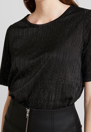 Vrouw draagt een zwart kort mouwtop met verticale glinsterende strepen en zwarte hoge taille broek met een rits aan de voorkant.