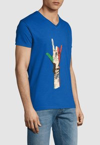 Camiseta de algodón azul con escote en forma de V, que presenta un gráfico de una mano en colores rojo, verde y beige, mostrando un signo de paz.