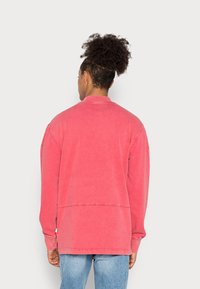 Mikina s dlouhým rukávem, vybledlá růžová barva, s kulatým výstřihem, spadlými rameny a volným střihem. Obsahuje viditelné švy a lem s vysokým a nízkým střihem.