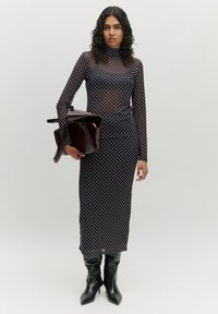 Schwarzes Netzkleid mit weißen Polka-Punkten, hohem Hals, langen Ärmeln und figurbetontem Schnitt. Das Model hält eine dunkelburgunderfarbene Handtasche und trägt kniehohe schwarze Stiefel.