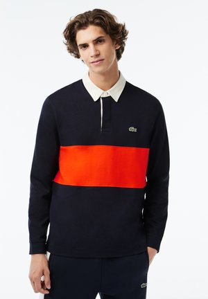 Camisa polo de manga larga de color azul marino con una franja central de color naranja brillante y un cuello de color claro en contraste; presenta un pequeño logo verde.