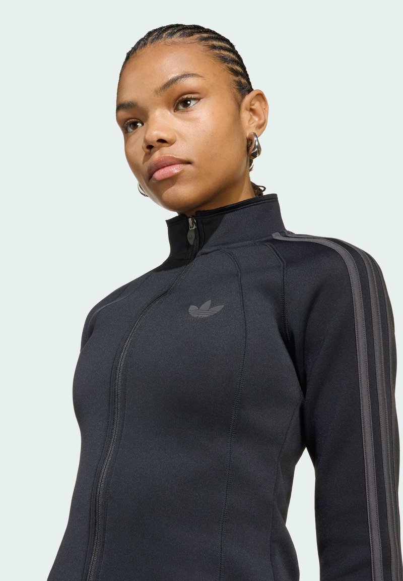 Kvinde med fletninger iført en sort Adidas lynlåstrøje med tre grå striber på ærmerne og et lille logo på brystet.