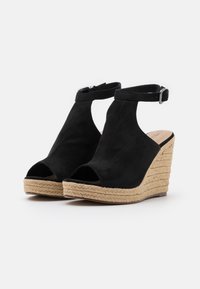 Černé semišové sandály na klínu s otevřenou špičkou, nártní páskem a juteovým obložením platformy. Vyznačují se hladkým povrchem a stříbrnou sponou.