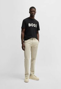 Sort T-shirt med hvid "BOSS" skrift, lys beige bukser og cremede sneakers. Glat stof, afslappet pasform og minimalistisk design.