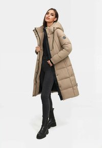 Abrigo largo acolchado beige con capucha, cremallera y botones a presión, con un diseño acolchado y bolsillos laterales, llevado sobre un top negro y jeans.