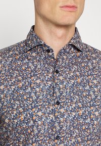 Close-up van een man in een marineblauw overhemd met knopen, versierd met een oranje en wit bloemmotief en met een open kraag.