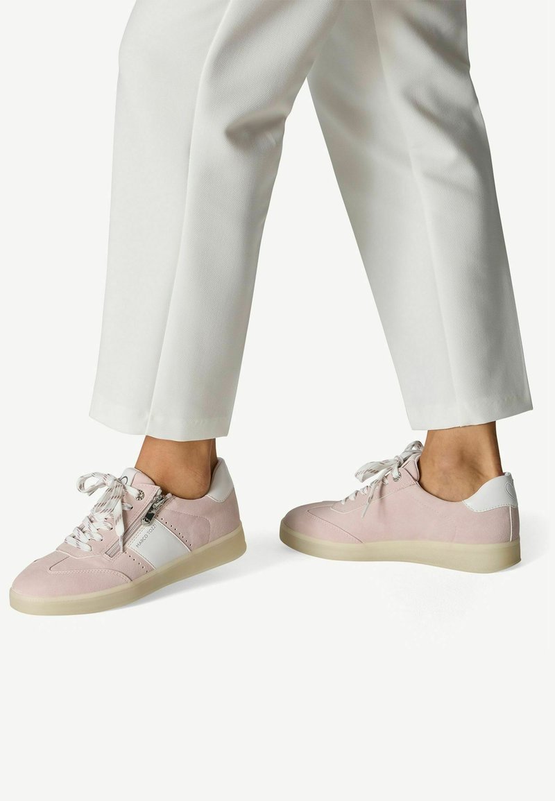 Zapatillas deportivas de ante rosa claro con detalles de cuero blanco, puntera redonda, cierre de cordones y suela texturizada, combinadas con pantalones blancos recortados.