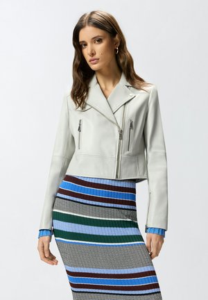 Žena u kratkoj svijetlosivoj kožnoj jakni sa patentnim zatvaračima i višebojnoj prugastoj pletenoj suknji, stoji ispred jednobojne pozadine.