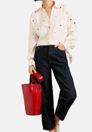 Vrouw draagt een wit overhemd met kleine rode en beige hartjes, donkere spijkerbroek, beige schoenen en houdt een glanzende rode shopper vast.