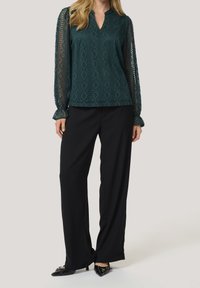 Blouse à motif bleu sarcelle avec un col en V et des manches longues transparentes, associée à un pantalon noir à jambes larges et des chaussures noires pointues.