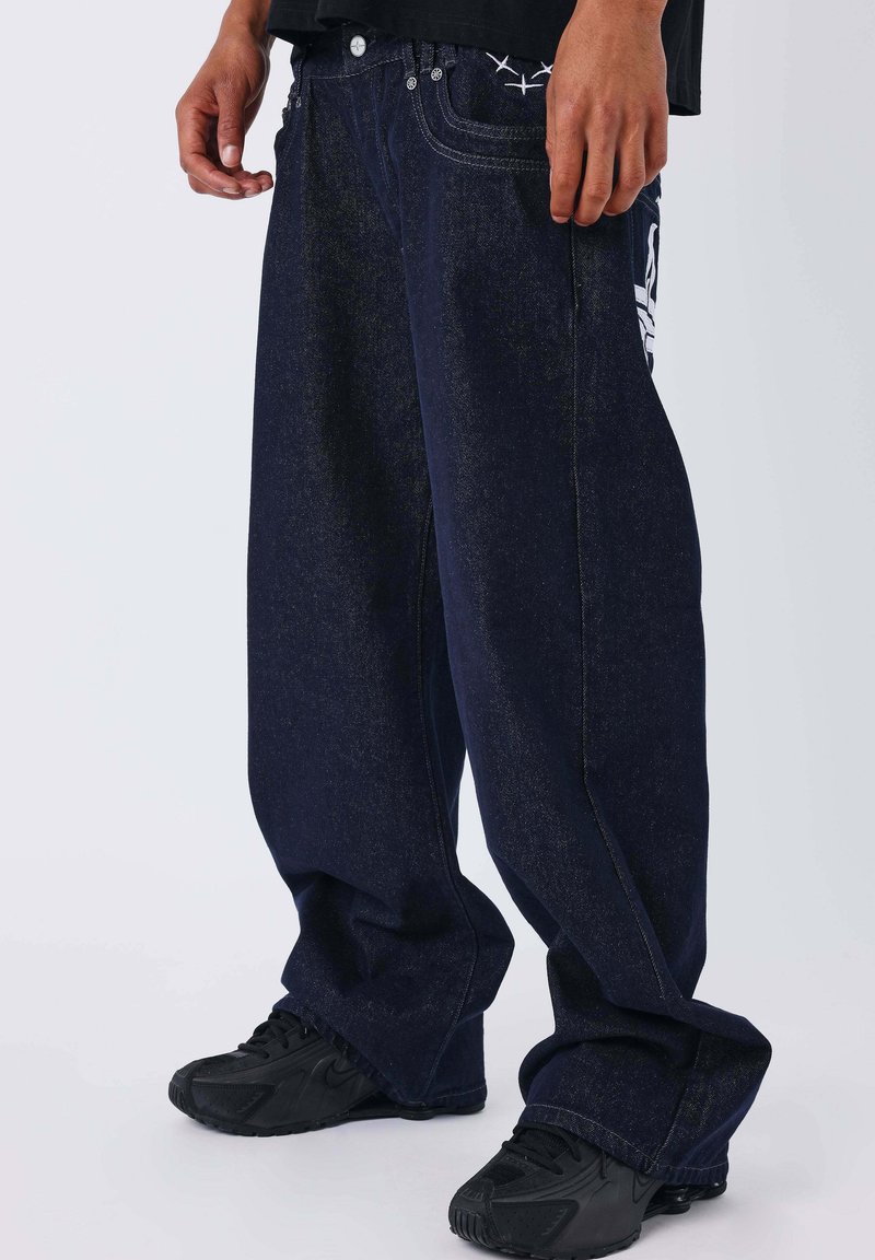 Jeans in denim blu navy con vestibilità ampia, caratterizzati da un ricamo bianco sulla tasca posteriore e da cuciture dorate a contrasto. Indossati con scarpe da ginnastica nere.