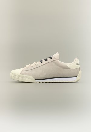 Low-top beige suede sneaker with white rubber sole, black and white laces, and branded heel tab on a neutral background.
