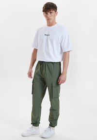 Hvid t-shirt med logo, parret med grønne cargobukser med sidelommer og elastiske manchetter; bæres med hvide sneakers.