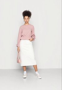 Blusa de manga longa rosa claro com punhos franzidos, combinada com uma saia branca até ao joelho e ténis brancos. Uma pequena mala rosa completa o visual.