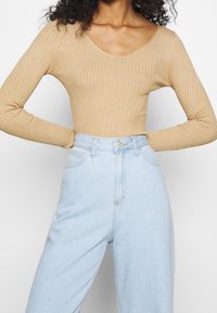 Kobieta w beżowej, prążkowanej bluzce z długim rękawem, włożonej w jasnoniebieskie jeansy z wysokim stanem i kieszeniami z przodu.