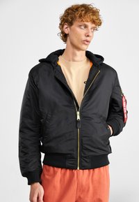Zwarte bomberjack met capuchon, gouden rits, geribbelde manchetten en zoom, oranje voering en een rode labeldetail op de mouw. Draagt over een beige shirt.