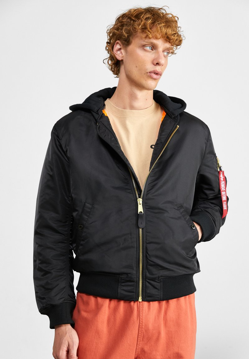 Zwarte bomberjack met capuchon, gouden rits, geribbelde manchetten en zoom, oranje voering en een rode labeldetail op de mouw. Draagt over een beige shirt.