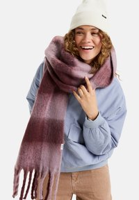 Oversized, zachte sjaal in gradient tinten paars met franjes. Casual gedragen met een lichtblauwe jas en een witte muts.