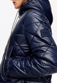 Námornícky pufový kabát s prešívaným dizajnom, kapucňou a šnúrkou. Obsahuje značkovú nášivku na rukáve a hladký, lesklý povrch.