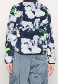 Miękka granatowa kurtka z białym i neonowozielonym kwiatowym wzorem. Długie rękawy, krótki fason i teksturowana tkanina. Charakteryzuje się kontrastowymi szwami.
