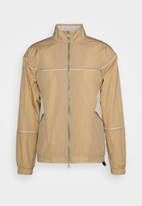 Ljusbeige vindjacka med hög krage, dragkedja fram, elastiska ärmmuddar och reflexvita detaljer längs axlarna och dragkedjorna.