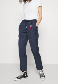 Námořnické kalhoty s pruhy, elastickým pasem a šňůrkou. Obsahují boční kapsy a malou vícolorovou nášivku. Nosit s černými teniskami.