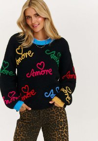Czarny sweter o dzianinowym splocie z kolorowym haftem tekstu "Amore" w odcieniach różu, zieleni, niebieskiego, żółtego i czerwonego; zestawiony z spodniami w panterkę.