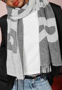 Sciarpa a motivi grigi e neri realizzata in materiale morbido, con bordi frangiati e un design moderno, indossata sopra una maglietta bianca e un blazer nero.