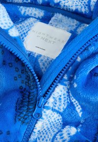 Jachetă din fleece albastră cu texturi și modele variate, dotată cu un fermoar albastru. Eticheta spune "NIGHTWEAR by NEXT," fabricată în China.