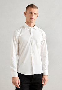 Hvit skjorte med knapper og myk tekstur, krage og lange ermer. Har en buet kant og subtile designdetaljer. Bært med mørke bukser.