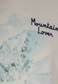 Grafica di montagne blu e verdi con testo ricamato nero "Amante della Montagna" su tessuto a maglia beige.