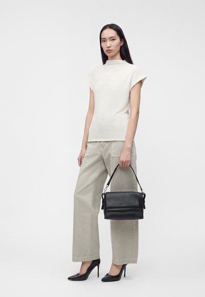 Femme portant un top blanc sans manches, un pantalon large clair, des talons hauts noirs, tenant un sac à main noir sur un fond uni.