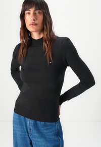 Maglione nero a maniche lunghe con collo a imbuto, dalla silhouette aderente e polsini a coste, abbinato a jeans blu a vita alta. Presente un dettaglio logo discreto.