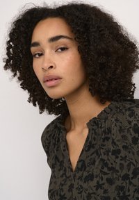 Blouse vert foncé avec un motif floral noir, tissu texturé et col à volants. Comprend une patte de boutonnage partielle à l'avant.