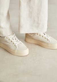 Helle beigefarbene Leinenschuhe mit einer dicken cremefarbenen Sohle, versehen mit goldfarbenen Ösen und weißen Schnürsenkeln, auf einem neutralen Hintergrund platziert.