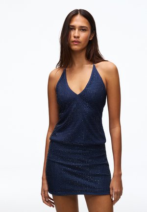 Femme aux longs cheveux foncés portant une mini-robe sans manches bleu marine scintillante avec un décolleté en V profond sur fond blanc.