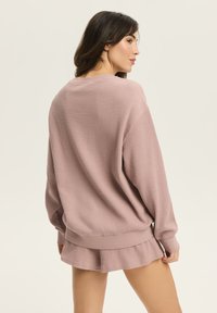 Sweter typu waffle w delikatnym różowym kolorze, z luźnym fasonem, długimi rękawami oraz ściąganym wykończeniem. W zestawie ze spódniczką o falbaniastym wykończeniu w tym samym kolorze.